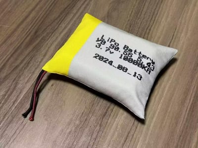 一张展示LiPo电池袋的照片，放置在木质表面上。电池袋呈长方形，左侧为黄色，右侧为白色，表面印有黑色文字，内容包括"LiPo Battery"、"3.7V"、"18000mAh"以及日期"2024_08_13"。电池袋一端伸出两根电线，分别为红色和黑色。