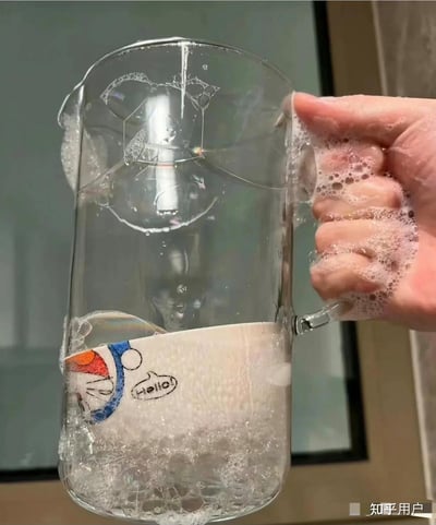 这张图片拍摄的是一个人在清洗带有哆啦A梦图案的玻璃杯，杯身上残留的肥皂泡在杯口处意外形成了类似Doge表情的狗头轮廓，充满了巧合和幽默感。细看泡沫构成的形状，可以清晰辨认出类似“Doge”经典表情的造型，令人会心一笑。

**巧合梗提示**：肥皂泡巧妙地勾勒出Doge狗头的脸型，属于典型的“错位梗”或“随机幽默”。