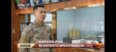 这是一张央视军事频道(CCTV 7)的新闻截图，画面中一名身着迷彩服的士兵正在接受采访。背景显示为一个现代化的食堂或餐饮设施，士兵表情严肃，正在谈论军队炊事工作。屏幕上有"新疆军区某旅 萨达姆"的文字标识和"我们的炊事员可以更专注于菜品的口味"的采访字幕。