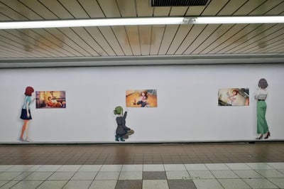 这是一张地铁站台的照片，画面中央是一面白色墙壁，上面展示着三幅动漫场景画作，以及三个动漫风格的人物剪影。左侧是红发女孩站立注视第一幅画，中间是绿发角色跪地伸手指向第二幅画，右侧是棕发女性站立观看第三幅画。整体场景给人一种文艺且治愈的氛围，仿佛动漫角色们也在欣赏艺术作品。