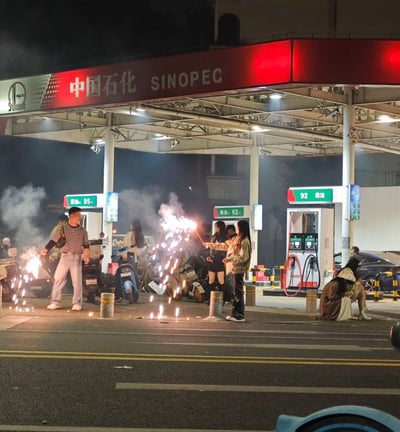 夜间的中国石化加油站场景，红色招牌上清晰显示“中国石化 SINOPEC”字样。站内有多个燃油加油机，分别标注“92 柴油”和“95 汽油”。数名人员在加油机附近燃放烟花，火花四溅，周围停放着电动车和一辆深色轿车。部分人站立手持烟花，另有两人坐在地上，现场存在明显安全隐患——加油站作为易燃易爆场所，严禁明火及燃放烟花爆竹。