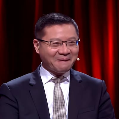 这是一张节目录制现场的视频截图，画面中的人物是学者张维为，他身着深色西装内搭白色衬衫，搭配格纹领带，佩戴黑框眼镜，耳侧装有领夹式麦克风，面带略带克制的温和笑容，背景是暗红色的幕布，整体场景正式，人物神情亲和，看起来正处于节目录制的环节中。