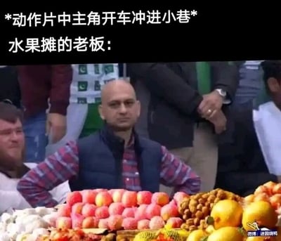 这是一张二次创作的搞笑梗图，采用文字加注的图文混排形式。上方用文字设定了经典动作片场景“动作片中主角开车冲进小巷”，下方是一位光头水果摊老板的照片，他双手叉腰，脸上带着无奈又生无可恋的表情，面前是摆满桃子、柠檬、山竹等各类水果的摊位。该梗图调侃了动作片里频繁出现的主角开车冲进小巷撞翻路边水果摊的俗套桥段，通过老板的表情生动表现出他面对这种“无妄之灾”时崩溃又无力的内心状态，十分贴合观众对这类桥段的吐槽心理。