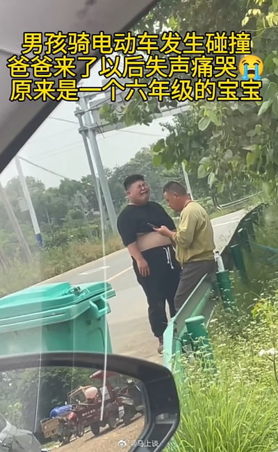 这张图是从汽车内向外拍摄的实拍视频截图，属于社会事件的传播素材。画面顶部配有黄色大字说明事件：男孩骑电动车发生碰撞，爸爸来了以后失声痛哭，原来是一个六年级的宝宝。画面中心身材壮硕的男孩穿着黑色短上衣和黑色长裤，正站在路边皱着脸委屈痛哭，上衣被撑起露出肚子；旁边的中年男性是男孩的父亲，正倚靠在路边的绿色护栏上低头查看手机，似乎在处理事故相关事宜。画面左下角可以看到汽车的后视镜，镜中反射出路边停放的三轮车等车辆，路边还有绿色垃圾桶、杂草树木以及交通电线杆，整体是郊外乡村道路的场景。
