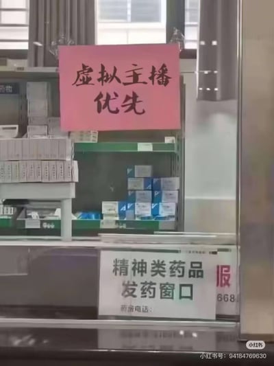 图片展示了一家药店的精神类药品发药窗口场景，玻璃窗上醒目地贴着一张粉红色手写标牌，上面用黑色毛笔字体写着“虚拟主播优先”。窗口内可见多层货架，整齐摆放着各类药品包装盒，背景为药店常规的灰色窗帘和白色窗框。标牌内容将“虚拟主播”这一互联网流行职业与严肃的药品发放场景结合，形成强烈的场景反差，带有幽默与讽刺意味，暗讽当下社会中某些领域盲目追逐网络潮流的荒诞现象。