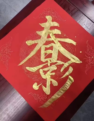这是一张正方形的红底书法斗方，以金墨手写了“春”与“樂（乐）”组成的合体字，背景印有精致的金色龙纹暗纹，笔触饱满有力，整体风格喜庆祥和，是专为春节创作的传统吉祥书法作品，适合作为新春期间的家居装饰或赠礼，传递新春欢乐的美好寓意。