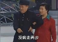 图片为经典小品《卖拐》的舞台截图，左侧穿黑色上衣的赵本山正搀扶着右侧穿红色上衣的范伟，两人站在带有几何图案的舞台背景前。赵本山表情严肃，身体微微前倾，范伟则面露无奈，配合着做出迈步的动作。图片底部配有白色字幕“没病走两步”，这是该小品中用来调侃假装腿脚不便者的标志性台词。