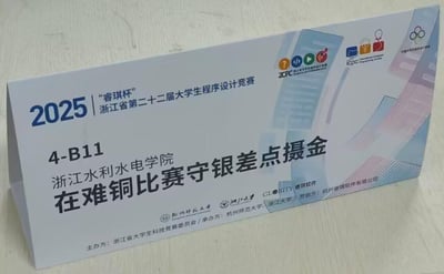 图片展示了2025年'睿琪杯'浙江省第二十二届大学生程序设计竞赛的指示牌。牌面顶部印有赛事全称及年份'2025'，中间标注了场地信息'4-B11 浙江水利水电学院'，并配有醒目文字'在难铜比赛守银差点摄金'（疑似为参赛队伍的趣味标语，可能存在谐音或笔误，原意为在比赛中努力争取更好成绩）。底部列明主办方为浙江省大学生科技竞赛委员会，承办方为杭州师范大学，协办方包括浙江大学，赞助方为杭州睿琪软件有限公司。右侧有多个赛事相关标识及ICPC等组织的logo，整体设计简洁，信息明确，用于竞赛场地指引。