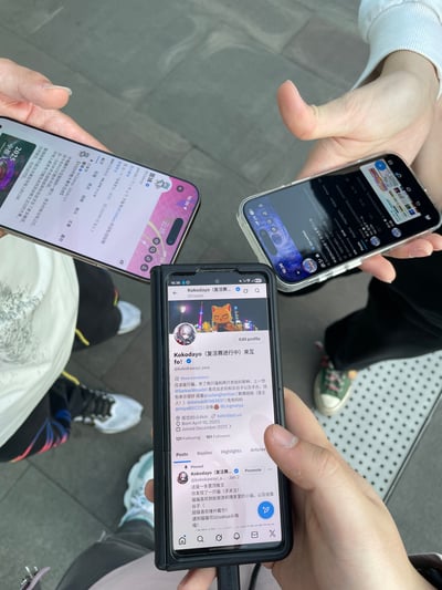 图片展示了面基现场的场景，三位人士手持智能手机，屏幕分别显示不同内容。中间手机屏幕清晰展示了社交账号“Kokodayo (魔法进行中) 菜五”的个人主页，包含猫形头像、简介文字（“已经是好朋友！扩列请私信我的助手哟...”）及关注/粉丝数据。左侧手机为粉色界面，右侧为深色界面，均显示文字内容。背景可见人物腿部及地面，推测为户外公共场所。