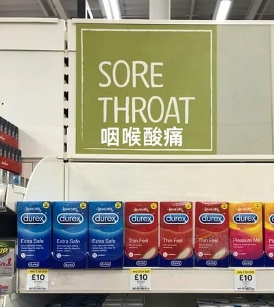 图片展示了超市货架场景，货架上整齐排列着多款杜蕾斯(Durex)避孕套产品，包括Extra Safe、Thin Feel、Pleasure Me等系列，每个包装价格均标注为10英镑。货架正上方悬挂着一块绿色标牌，上面用白色文字醒目地写着英文“SORE THROAT”和中文“咽喉酸痛”，形成产品功能与标牌指示之间的幽默错位对比，引发观者对两者关联性的趣味联想。画面清晰呈现了超市货架结构、产品包装细节及价格标签，整体场景真实自然。