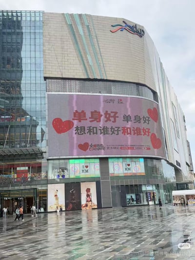 这是一张雨后实拍的昆明同德昆明广场户外场景图，广场地面湿漉漉的，商场外的大型弧形LED屏上展示着趣味单身主题标语：“单身好 单身好 想和谁好和谁好”，搭配多个红色爱心图案，下方还有“全糖热恋 心动企划”及“更多精彩 小红书搜索 同德昆明广场”的小字。该标语以幽默接地气的口吻调侃单身状态，打破传统情侣向节日营销的常规，引发单身群体共鸣，是商场的创意营销内容。
