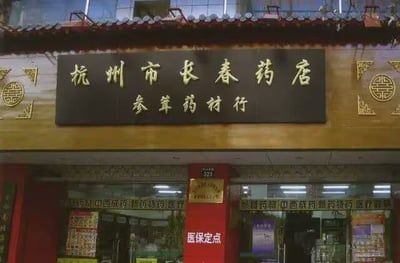 这是一张实体药店的门头实拍图，整体采用中式风格设计：门头上方悬挂深棕色底金色字体的招牌，主文字为“杭州市长春药店”，下方小字标注“参茸药材行”，招牌两侧配有金色的中式寿字纹和角纹装饰；招牌下方的红色立柱上印有门牌号、金色荣誉牌匾以及白色的“医保定点”字样；门店为玻璃门脸，玻璃上印有经营零售药材、中西成药、特药、医疗器械等品类的相关文字，可隐约看到店内的灯光与货架陈设。该图因店铺名称“长春”谐音“常存”，结合药店属性，被网友关联到囤药相关的热点话题进行传播。