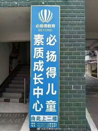 图片展示了一个蓝色的教育机构招牌，位于一栋建筑的入口处。招牌上方有“必扬得教育 BEYOND”的标志和文字，中间竖排大字为“必扬得儿童素质成长中心”，底部标注“由此上二楼”及联系电话（部分号码模糊）。背景是绿色砖墙，左侧可见楼梯入口，整体为真实场景的实拍照片。