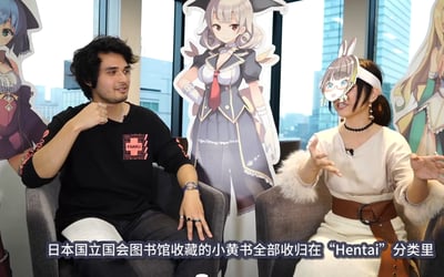 这是一段访谈类视频的截图，画面呈现室内访谈场景：一男一女分别坐在灰色沙发上交流，背景立着多幅二次元美少女角色立牌，窗外可见城市高楼景观。左侧男士身着印有“FAKKU”字样的黑色上衣，面带微笑侧头看向对方，左手抬起做出手势；右侧女士戴着兔子耳朵造型的搞怪头箍，面部有遮挡，双手摊开，正生动地表达观点。画面底部配有白色字幕，内容为：“日本国立国会图书馆收藏的小黄书全部收归在“Hentai”分类里”，整体氛围轻松诙谐，围绕二次元成人文化相关话题展开讨论。
