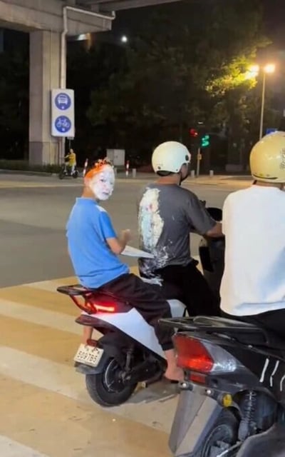 夜晚的街道十字路口，三个人骑着两辆电动车等待通行。穿蓝色T恤的男孩脸上涂满白色和橙色颜料，疑似刚参加完化妆活动；中间穿灰色T恤的骑手后背有大面积彩色涂鸦，两人均佩戴头盔（白色和黄色），右侧穿白色T恤的骑手同样戴着黄色头盔。背景可见自行车道指示牌、绿色交通信号灯及远处骑电动车的路人，整体画面充满生活气息又略带荒诞感。