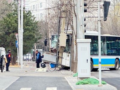 一张城市街道交通事故现场的照片。画面中央可见一辆蓝白相间的公交车前端严重受损，撞上了路边的电线杆，导致公交车挡风玻璃碎裂，车头部分结构变形。电线杆也明显倾斜，周围散落着一些碎片和设备。两名身穿深色外套的男子站在事故现场附近，其中一人似乎正在查看情况或使用手机。背景中有光秃秃的树木、交通信号灯、路灯和一些建筑物，显示这是一个城市街道环境。整体场景给人一种突发事故后的紧张和混乱感。