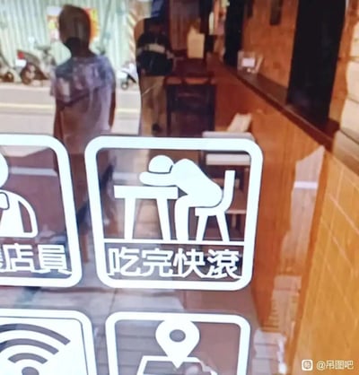 这是一张线下店铺玻璃的实拍图，玻璃表面贴有多款图标式标识，其中最具话题性的是一个图案为人物从餐桌前起身的标识，下方配文“吃完快滚”，用戏谑幽默的方式调侃式地催促顾客用餐后尽快离开，以此提高店铺翻台率；旁边还配有“店員”标识、WiFi标识等。背景中可以看到店铺内部的陈设，以及店外街道上站立的行人，整体画面充满趣味，将商家的诉求用夸张搞笑的标语呈现出来，属于网络上流行的搞笑店铺文化内容。
