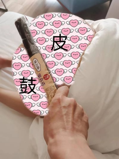一张创意搞笑的食物梗图，画面中一只手拿着一包传统零食'果丹皮'，背景是白色的被子。图片中央有一个粉色爱心图案的圆形覆盖层，上面用黑色大字写着'皮'和'鼓'两个汉字，巧妙组成'皮鼓'(pí gǔ)的谐音梗，与零食名称'果丹皮'中的'皮'字呼应，形成有趣的视觉文字游戏。