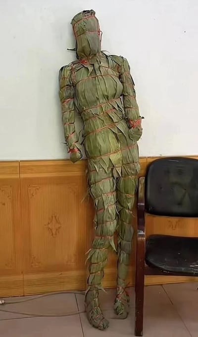 室内场景中，一个真人大小的人形被绿色粽叶层层包裹，用红色细绳横向捆绑固定，呈现出类似粽子的纹理结构。人形靠墙站立，姿态自然，头部、躯干、四肢均由粽叶覆盖，细节处可见粽叶的自然褶皱。背景为白色墙面，下方是带有雕花图案的木质柜子，右侧放置一把黑色塑料椅子，整体环境像是教室或办公室。该造型因使用端午节标志性的粽叶材料，充满节日创意感，被网友戏称为“端午节限定人形粽子”。