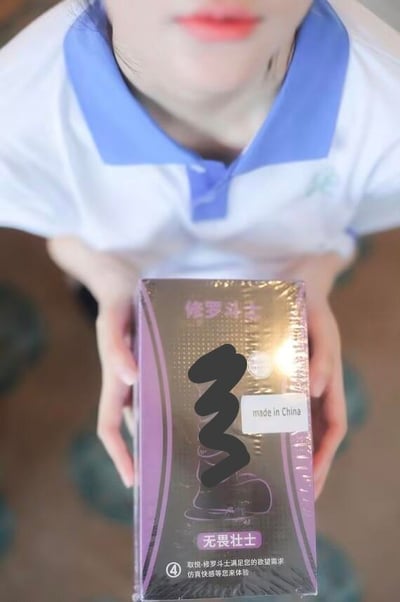 图片展示了一个身穿白色上衣配蓝色领子服装的人，双手捧着一个金色和紫色相间的包装盒。盒子上印有“修罗斗士”和“无畏壮士”等字样，并有“made in China”的标签。盒子部分区域有黑色涂鸦遮盖。整体拍摄角度为从上往下，突出展示人物手中的产品。