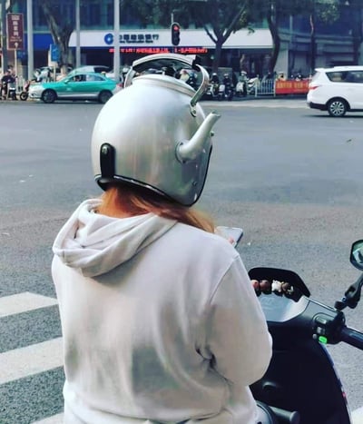 在城市的十字路口，一位骑电动车的女士背对着镜头，她戴着一个由不锈钢水壶改造而成的创意头盔，水壶的壶身、壶嘴和提手都完整保留，造型独特又搞怪，瞬间成为视觉焦点。女士身穿浅灰色连帽卫衣，正手持手机，似乎在等待红灯。背景中可以看到城市街道、过往车辆以及中国建设银行的网点，充满日常的生活气息。这张图没有双关或谐音梗，完全是靠新奇的改造创意带来趣味效果。