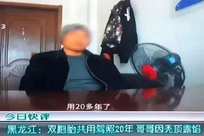 这是一则社会新闻的视频截图，画面中一位头部被马赛克处理的老年男性坐在桌前，处于室内环境中，背景可见房门、挂着的外套和风扇。图中字幕和人物话语显示，黑龙江的这对双胞胎兄弟共用一本驾照长达20年，最终因为哥哥秃顶，与驾照上的照片产生明显差异，导致这件事败露，老人在画面中说道“用20多年了”。