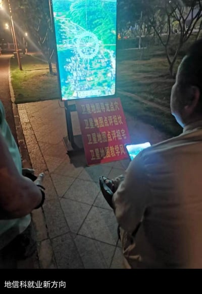 夜晚户外的人行道边，一个摆摊的摊位立着显示屏，展示带有风水罗盘标记的卫星地图，下方红色招牌标注着“卫星地图寻龙点穴、点评祖坟祖宅、教学风水”的业务内容，有路人在旁围观。图片底部配文“地信科就业新方向”，用地理信息科学专业的核心技能——卫星地图运用，结合传统风水业务玩梗，调侃地理信息类专业的另类就业思路，通过专业技能和传统行业的反差结合制造搞笑效果。