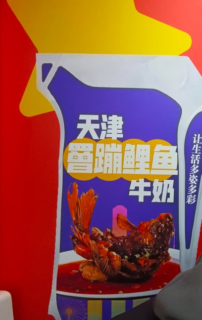 这是一张趣味创意宣传海报，采用牛奶包装的造型设计，将天津特色名菜罾蹦鲤鱼与牛奶进行创意结合。海报主体区域为紫色背景，上方醒目标注“天津 罾蹦鲤鱼 牛奶”，下方展示了色泽浓郁、造型完整的罾蹦鲤鱼菜品图，右侧配有标语“让生活多姿多彩”，整体风格活泼亮眼，主打地域特色美食与乳品的新奇混搭，带来有趣的视觉冲击。