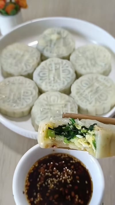图片中展示了一盘外形酷似月饼的白色圆形点心，表面带有传统月饼的花纹。其中一块被筷子夹起，露出内部翠绿的蔬菜和黄色的鸡蛋馅料，看起来像饺子馅。旁边还有一小碗撒了芝麻的深色蘸料。这种食物外形像月饼，内馅似饺子，让人傻傻分不清到底是月饼、饺子还是汤圆，充满了趣味性。