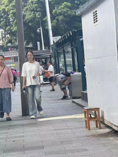 这是一张城市街头的实拍照片，画面中一名男子弯腰在垃圾桶旁，由于视觉错位，他的上衣和身体角度让他看起来像是脱下裤子露出臀部，实际是视觉错觉造成的趣味效果。
没有双关、谐音梗或暗示内容，纯粹是巧合形成的视觉错位。