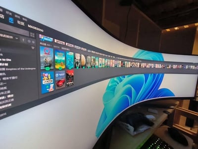 这是一张实拍的超宽曲面显示器画面，屏幕上呈现Steam客户端的游戏库界面，左侧是游戏列表，中间及上方排列着大量游戏封面，包括《战神》《星露谷物语》《VRCHAT》等知名游戏，背景是Windows系统的默认蓝色壁纸。显示器的曲面形态让画面呈现出独特的弧形视觉效果，直观展示了大屏设备在游戏内容浏览上的宽阔视野，给人震撼的视觉体验。