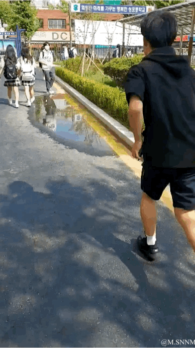 这是一段动态GIF，画面呈现户外街头场景：一名身穿黑色短袖短裤的男生，悄悄跑到路边的积水区域旁，做出要抬脚溅水的动作，目标是前方步道上行走的几名女学生，周围有绿化带、店铺和韩文标识，整体是充满恶作剧趣味的日常瞬间。