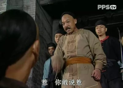 这是一张清代古装影视剧的截图，画面核心是一位身着浅棕色清代服饰的中年官员，他站在古旧砖墙门洞前，表情愤怒，手指着对面的人物，口中怒喊“惹 你敢说惹”，身后跟随着几位穿着深色服饰的随从，整体场景氛围紧张，传递出强烈的冲突感，官员的愤怒情绪十分突出，似乎是被对方的言语彻底激怒。