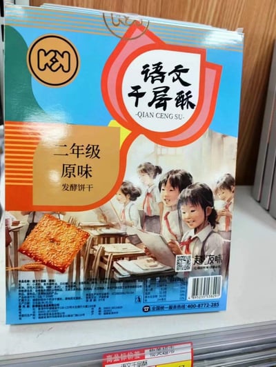 这是一张展示名为'语文千层酥'的发酵饼干包装的照片。包装主体为蓝色，顶部有红色盾形装饰，中央印有产品名称'语文千层酥'及拼音'QIAN CENG SU'，左侧橙色区域标注'二年级 原味'字样。包装正面有一幅插画，描绘了几位穿着校服、系着红领巾的小学生围坐在一起阅读的场景，背景为教室环境。右下角有品牌名'超友味'及二维码，底部印有营养成分表和条形码。整体设计结合教育元素与食品包装，通过'语文'学科名称和'二年级'年级标注营造怀旧校园氛围。