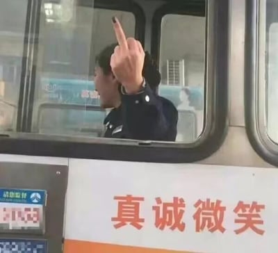 这是一张原始拍摄的静态图，画面呈现的是一个公共服务窗口，从场景和工作人员制服判断，可能是高速公路收费站或城市公交服务窗口，窗口内的工作人员穿着深色制服，侧脸朝向画面外侧，一只手伸出窗口，做出了竖中指的不礼貌手势。而在窗口下方的白色墙体上，用醒目的红色大字印着“真诚微笑”的服务标语，工作人员的负面行为与下方倡导文明服务的标语形成了极其强烈的反差，带有明显的讽刺意味，凸显了服务标语与实际服务态度的脱节。图片左下角还有一个蓝底白字的“请您监督”的标识，进一步加剧了这种讽刺效果。