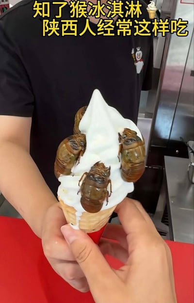 这是一张网友实拍的整活趣味图片，画面前景有一双手捧着一只甜筒冰淇淋，白色奶油部分插了四只知了猴（金蝉若虫，是部分地区流行的食用昆虫），背景可见一名穿着印有蜜雪冰城雪王标识黑色T恤的工作人员的上半身，图片顶部配有黄色醒目文字“知了猴冰淇淋🍦陕西人经常这样吃”，该内容为网友虚构的猎奇食物搭配，并非陕西地区真实存在的普遍吃法。