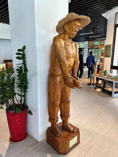 室内展厅中，一座木雕农夫雕像靠墙而立，雕像头戴草帽，上身赤裸，双手交握于身前，神情严肃。雕像旁有一盆红色花盆的绿植，背景可见其他参观者和展厅陈设，整体呈现出写实的艺术风格。