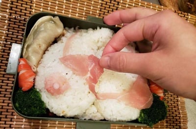 一张俯拍视角的美食照片，展示了一个绿色方形便当盒里的创意熊猫造型米饭。米饭被塑造成熊猫头部形状，一只手正用镊子（或手指）小心翼翼地将粉红色火腿片放在米饭上，作为熊猫的耳朵。便当盒内还配有其他食材：左侧有一个煎饺、橙色胡萝卜花片，底部铺有绿色西兰花，整体色彩搭配丰富，造型可爱。背景是竹制餐垫，增添了日式料理的氛围。