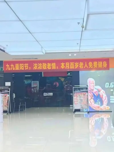 这是一张线下商场内健身房门店的实拍照片，门店入口上方悬挂着红色的重阳节主题宣传横幅，文字内容为“九九重阳节，浓浓敬老情，本月百岁老人免费健身”。门店旁的墙面上印有肌肉健美运动员的宣传海报，地面为光滑亮面瓷砖，可以倒映出海报的影像。该促销活动针对几乎没有健身消费需求、也很难具备健身能力的百岁老人推出免费福利，属于没有实际落地意义的营销噱头，充满荒诞的搞笑感。