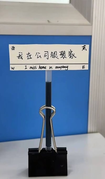 图片中展示了一张白色手写标语，被黑色长尾夹竖立固定在白色桌面上。标语中央用黑色字迹写着中文“我在公司很想家”，下方对应英文翻译“I miss home in company”，左右两端分别标注“西”（W）和“东”（E）。背景为蓝色柜子和浅色墙壁，整体呈现出办公环境中的一个小场景，幽默又真实地表达了职场人在工作时思念家乡的普遍情绪。