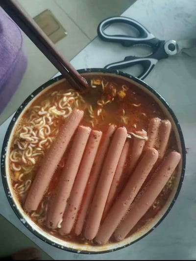 这是一张俯视视角拍摄的食物照片，画面中心是一碗盛在带黑边的搪瓷碗里的红汤泡面，泡面汤汁中整整齐齐码放了十根完整的火腿肠，占据了大半个碗的空间，一双深棕色筷子插在泡面左上角的汤汁里，碗外的大理石台面上还放了一把黑白色手柄的剪刀，画面左上角露出一小块紫色衣物，整份餐食火腿肠的量远超出常规泡面搭配，显得十分夸张。