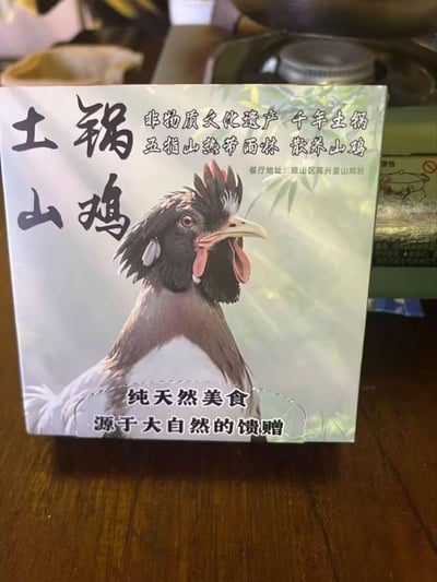 这是一张土锅山鸡美食包装盒的实拍图，盒面以一只神态昂扬的散养山鸡为核心视觉元素，搭配多组宣传文字：标注了“非物质文化遗产 千年土锅”“五指山热带雨林 散养山鸡”的产品背景，给出餐厅地址“琼山区高兴里山鸡坊”，并以“纯天然美食 源于大自然的馈赠”强调食材的原生态属性，整体风格质朴自然，主打海南原生态特色美食的卖点，无谐音梗、双关等特殊内容。