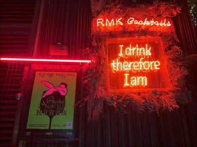 夜晚的酒吧 exterior，上方是红色霓虹灯招牌“RMK Cocktails”，下方是黄色霓虹灯标语“I drink therefore I am”（戏仿笛卡尔“我思故我在”的幽默改编），招牌周围装饰有绿色植物。左侧有一张黄色海报，上面有粉色图案和中文文字“一只一杯，深[内心]提的毛”“OneShot Night 独立宣言夜”，以及“LOVE U.B”“asOa”“RMK”等品牌标识，整体营造出时尚、轻松的夜生活氛围。