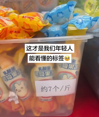 这张图展示了一个盛放乳酸菌可吸果冻的透明塑料盒，盒子上方还堆放了其他包装的零食，盒身贴有一张手写白色标签，标注“约7个/斤”，直观告知消费者该款果冻每斤的大致数量。图上叠加了两行配文“这才是我们年轻人能看懂的标签🥺”，吐槽日常很多散称商品仅标注每斤单价，消费者很难快速估算单个商品的价格，这种直接标注每斤对应个数的标价方式更简单易懂，契合年轻人的消费习惯。