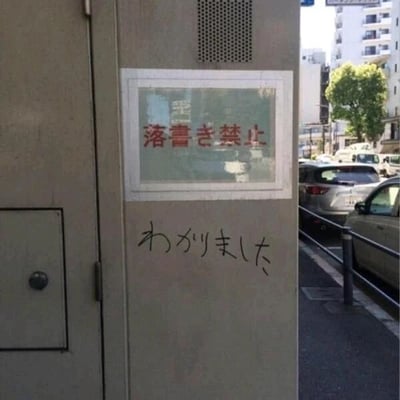 这是一张拍摄于日本街头的原始实拍图，画面主体是一个户外金属设备的墙面，墙面上贴着一个带有白色边框的标牌，标牌内用红色日文标注着“落書き禁止”，翻译为中文是“禁止涂鸦”。但在该标牌正下方的墙面上，有人用黑色笔迹手写了日文“わかりました”，翻译为中文是“我知道了”。这种行为形成了强烈的反差感：涂鸦者明明看到了禁止涂鸦的提示，却用涂鸦的方式回应自己已知晓禁令，用一种直白又戏谑的方式打破规则，制造出荒诞搞笑的幽默效果，属于典型的街头整活行为。背景中可以看到日本城市街道的景象，有停放的车辆和楼房，进一步佐证了这是街头场景。