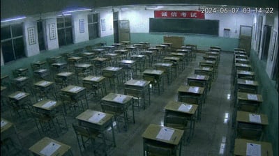 这是一张拍摄于2024年6月7日凌晨3点14分22秒的考场监控截图，6月7日为中国普通高等学校招生全国统一考试的开考首日，此时教室内已经完成考场布置：整齐排列的课桌上分别摆放了考试相关纸张和文具，教室前方黑板上方悬挂有红色的“诚信考试”标语，墙面还张贴有规章制度类张贴物，此时窗外为黑夜，教室内亮着照明灯光，空无一人，处于等待考生入场的考前准备状态。
