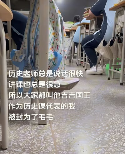 教室内部场景，学生们坐在排列整齐的课桌前，地面可见彩色图案的书包和白色运动鞋，后方黑板隐约可见。图片中央覆盖白色文字：“历史老师总是说话很快 讲课也总是很急 所以大家都叫他吉吉国王 作为历史课代表的我 被封为了毛毛”。这里巧妙借用动画《马达加斯加》中朱利安国王（吉吉国王）急躁多言的性格特点，将说话快的历史老师比作吉吉国王，而课代表则对应其助手莫里斯（毛毛），形成校园场景下的趣味角色联想。