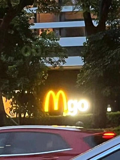 夜晚的户外场景中，树木枝叶遮挡了部分招牌，让麦当劳标志性的金色‘M’标志与后方亮着的‘go’字样恰好组合在一起，形成了看起来像‘Mgo’的视觉错觉效果。画面下方可见停放的车辆，背景是带有灯光的建筑物，整体氛围轻松有趣。