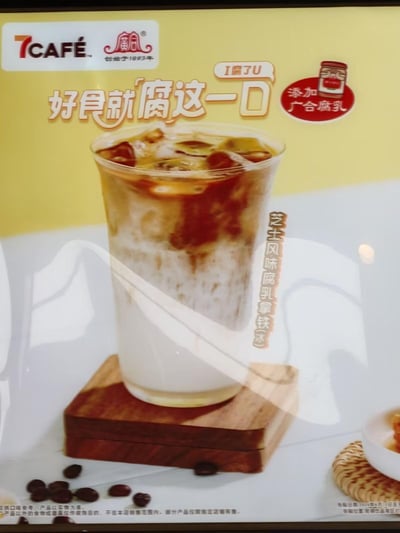 7CAFE与广合腐乳联名推出的芝士风味腐乳拿铁冰广告。画面主体为一杯分层明显的冰拿铁，杯中有冰块、咖啡与奶泡层，放置在木质杯垫上，周围散落咖啡豆。背景为黄色，上方有7CAFE和创立于1993年的广合腐乳品牌标识。主要宣传语包括“好食就腐这一口”“I腐了U”（谐音“我爱你”），并标注“添加广合腐乳”。饮品名称“芝士风味腐乳拿铁冰”竖排显示在杯身右侧。底部小字注明“仅作口味参考，产品以实物为准。产品以外的食物或餐具仅作装饰目的，不在本品销售范围内，部分产品仅限指定店铺有售。”