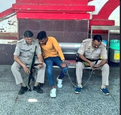 画面呈现了充满反差感的一幕：两名印度执法人员与一名被铁链手铐锁住的男子同坐在公共区域的长椅上。左侧执法人员手持步枪，却和中间的被看押男子一同低头专注看手机；右侧执法人员腰间配枪，自己也低头玩着手机，男子的手铐铁链还连接在他身上。本该严肃的执法看押场景，变成了三人一起摸鱼的画面，滑稽感拉满。